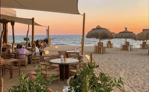 Terrasse du restaurant et plage du Assaona Beach Club à Portixol, Majorque, au coucher du soleil avec des dîneurs attablés sous des parasols en paille.