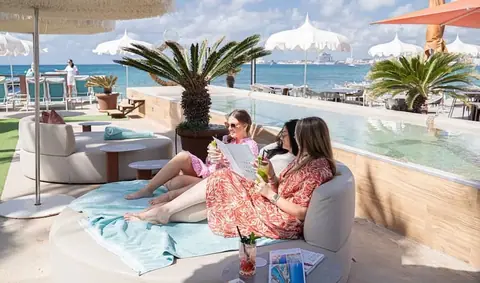 Groupe d'amies célébrant un EVJF sur un lit balinais au Anima Beach Club à Palma de Majorque, cocktails à la main, avec vue sur la piscine et la mer.
