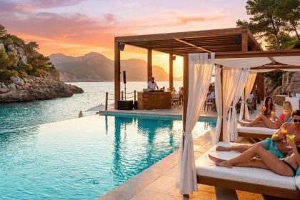 Beach club de luxe à Majorque au coucher du soleil, avec piscine à débordement surplombant une crique, clients sirotant des cocktails sur des lits balinais et DJ sous une pergola.