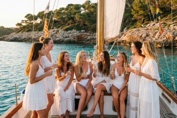 Groupe d'amies en tenue blanche célébrant un EVJF sur un voilier à Majorque, champagne à la main, au coucher du soleil.