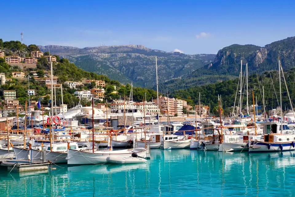 Vue panoramique du Port de Sóller à Majorque, avec de nombreux bateaux de plaisance amarrés dans le port naturel, la ville en bord de mer et la Serra de Tramuntana en arrière-plan.