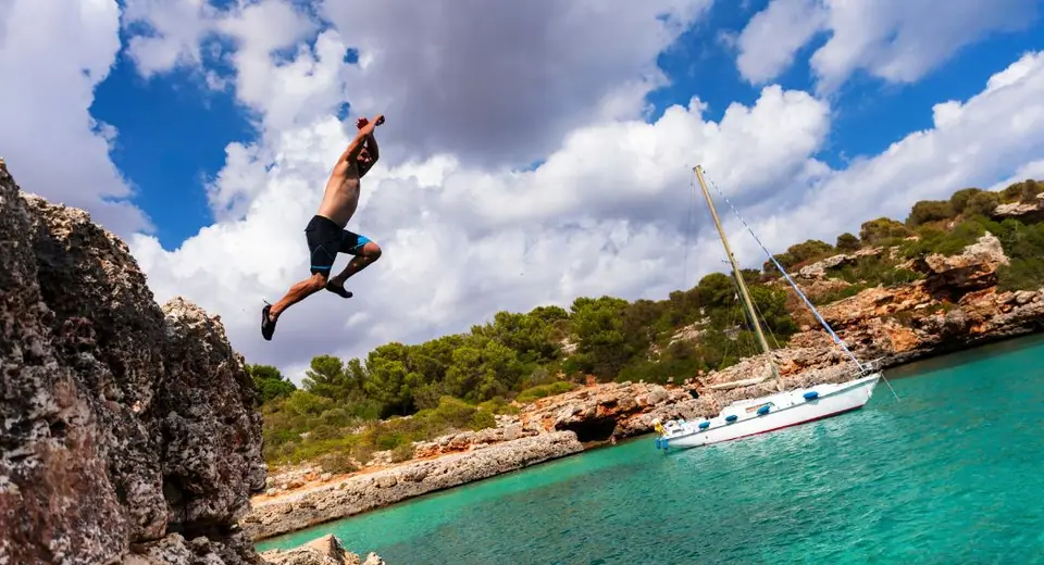 Homme sautant d’une falaise dans une eau turquoise à Cala Sa Nau à Majorque avec un voilier en arrière-plan